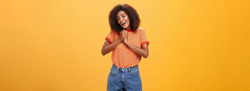 Girl sighing and gazing at cute thing clenching palms together near heart smiling with admiration and love being feminine and tender posing over orange background feeling affection. Copy space