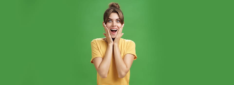 Indoor shot of excited and surprised energized attractive glamorous female in yellow t-shirt touching cheeks from amazement smiling broadly astonished posing over green background. Emotions concept
