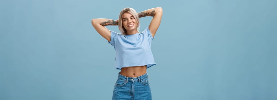 Thanks god it weekends. Relaxed lazy and attractive young stylish woman with fari hair tattoos, pierced belly stretching with delight holding hands behind head in carefree pose, smiling over blue wall.