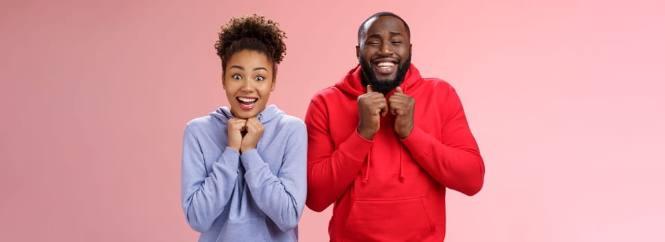 Excited happy girlfriend boyfriend smiling broadly nervously clenching fists pressed chest looking camera thrilled anticipate good news trying luck playing lottery together, standing pink background.