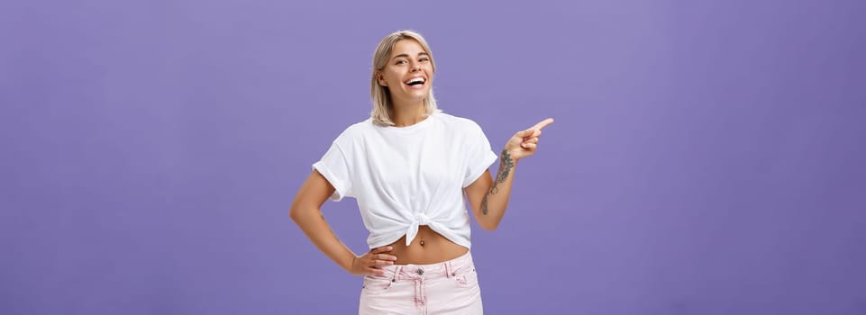 Check out cool place. Satisfied carefree stylish and attractive adult woman with perfect body tanned skin, tattoos laughing and smiling joyfully holding hand on hip and pointing left over purple wall.