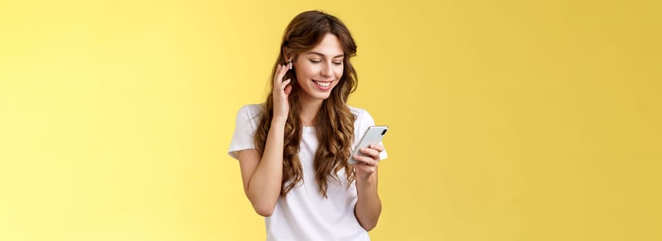 Tender feminine curly-haired female white t-shirt touch wireless earbud put earphone ear smiling pleased look smartphone screen pick song wanna listen music search right track yellow background. Lifestyle.