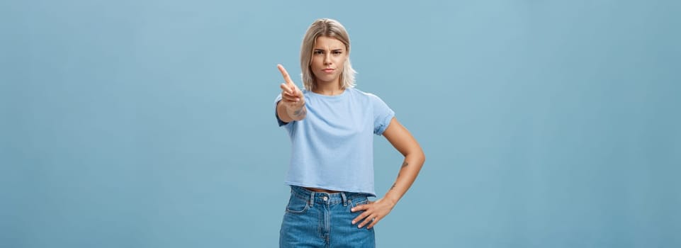 You better not do it. Serious-looking displeased strict elder sister with fair hair and tanned skin frowning holding hand on waist shaking index finger in forbid or stop gesture over blue background.