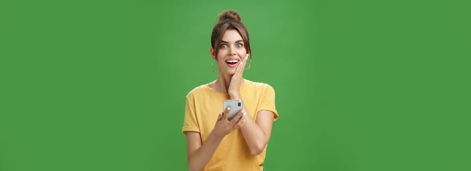 Surprised touched and impressed beautiful european girl with diastema smiling broadly touching cheek from amazement and joy holding smartphone looking happy at camera after reading cool news. Technology concept