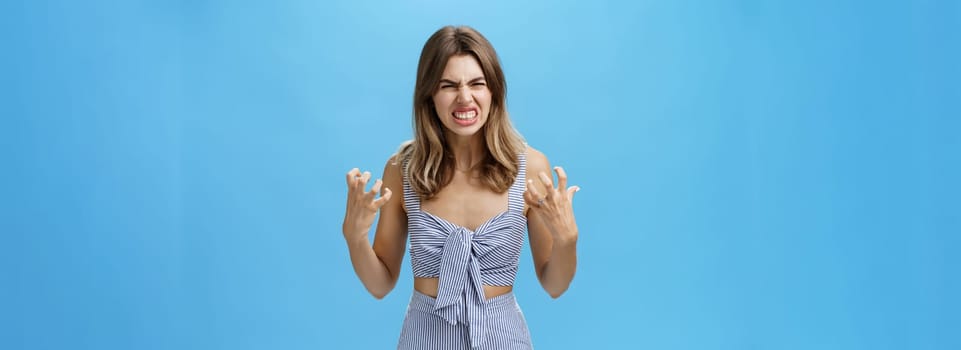 Indoor shot of irritated moody woman having mental breakdown feeling annoyed and pressured squeezing hands in fists from anger wrinkling nose in hate standing hateful over blue background. Emotions concept