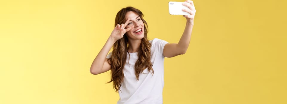 Joyful charismatic good-looking curly-haired woman white t-shirt show peace victory sign smiling broadly posing tender friendly expression smartphone front camera take selfie yellow background. Lifestyle.