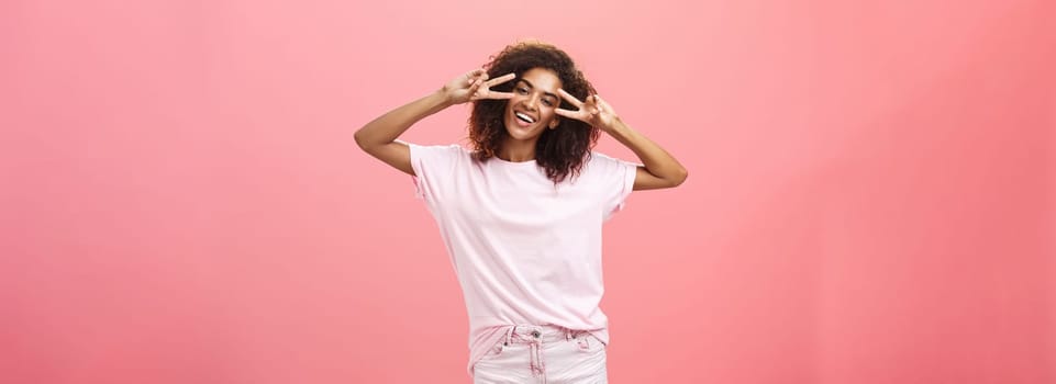 Indoor shot of charismatic playful dark-skinned woman with afro hairstyle tilting head showing peace or victory signs over eyes and smiling feeling amused chilling on awesome party over pink wall. Lifestyle.