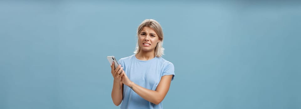Woman cannot stand when mom shouts during phone call. Intense displeased young european blonde with tanned skin clenching teeth and frowning covering microphone of smartphone over blue wall.