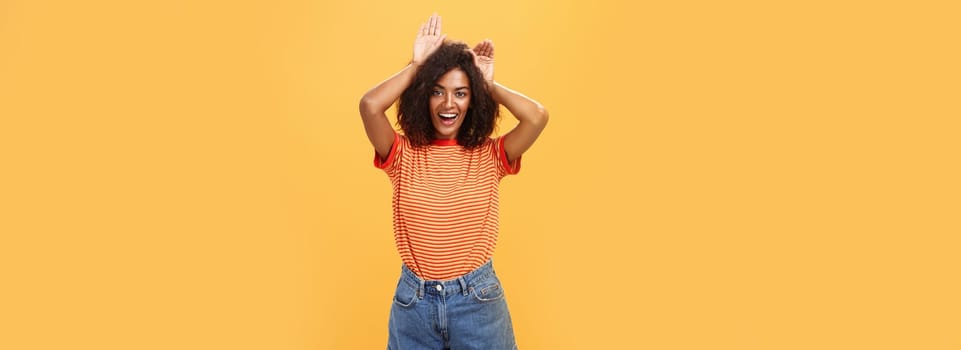 Let me be your bunny. Portrait of charming enthusiastic and charismatic happy dark-skinned female with afro hairstyle holding palms on head like animal ears making cute face over orange background.