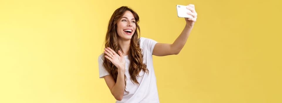 Friendly lively good-looking cheerful feminine girl extend arm hold smartphone record video blog waving front camera smiling broadly talking fans videocalling taking selfie yellow background. Lifestyle.