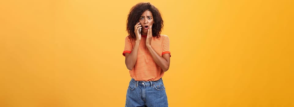 Woman receiving disturbing call feeling emapthy and sorry for poor friend getting in trouble holding cellphone near ear touching cheek concerned frowning standing sad, worried over orange background.