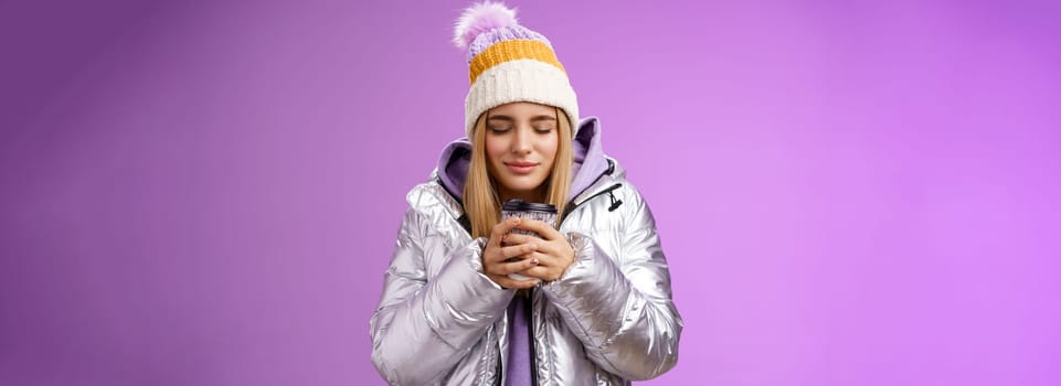 Charming tender romantic blond girl sniffing nice smell warm tasty coffee holding take-away cup warm hands close eyes look delighted wearing winter jacket hat enjoy vacation, purple background.