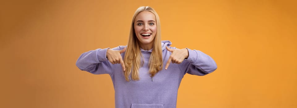 Amused joyful nice blond girlfriend pointing down present cool new product smiling broadly recommending try check out standing orange background happily grinning in hoodie. Advertisement concept
