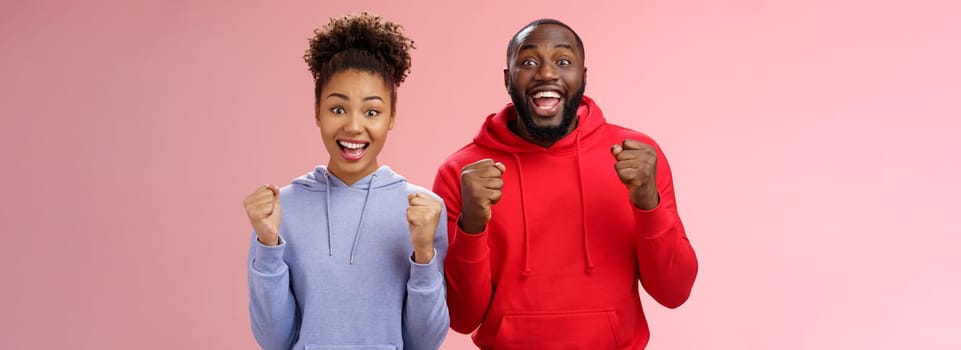 Lucky cheerful two african-american man woman yelling hooray celebrating triumphing huge success clenching fists joyfully accomplish mutual goal standing joyfully pink background victory gesture.