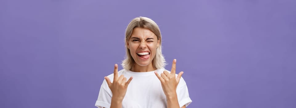 Gonna rock. Portrait of joyful happy good-looking stylish woman with blond medium haircut winking smiling and sticking out tongue while showing rock-n-roll gesture with both hands over purple wall.