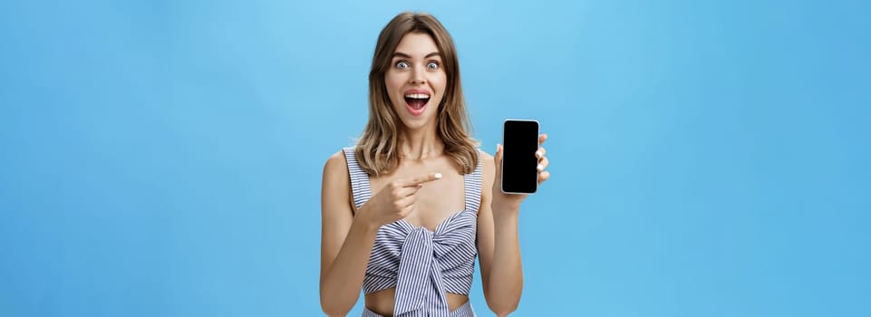 Excited happy woman with gapped teeth finally bought brand new smartphone holding device in hand pointing at cellphone screen showing cool app smiling broadly from joy against blue wall. Advertisement concept