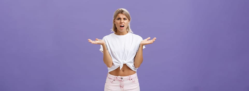 Lifestyle. Perplexed and disappointment good-looking blonde female student in white t-shirt and pink shorts frowning shrugging with spread hands near shoulders saying what the hell over purple wall.