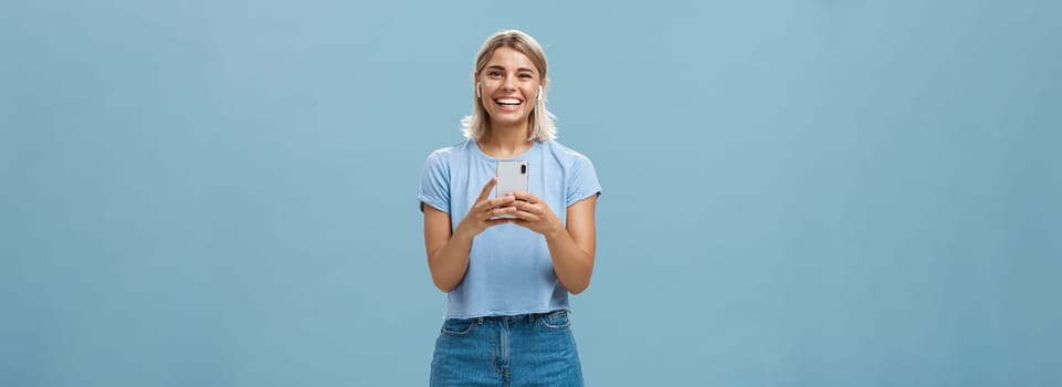 Satisfied happy and carefree young attractive blonde woman in casual outfit smiling broadly from happiness and delight listening music in wireless earphones holding smartphone over blue wall. Technology and lifestyle concept