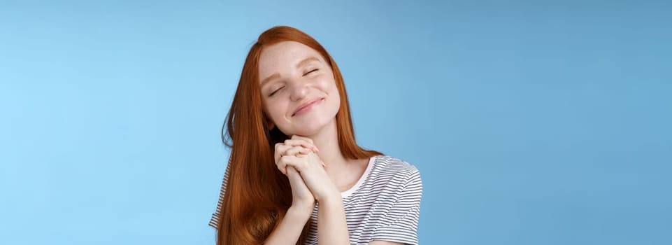 Charming delighted happy dreamy redhead girlfriend ginger long hair tilting head close eyes smiling pleased daydreaming thinking about sweet tender memories press palms together, blue background.