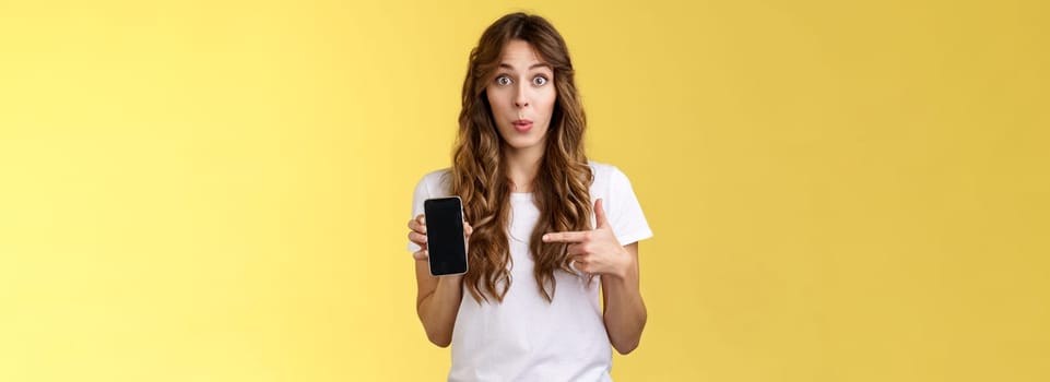 Intriguing app check out. Enthusiastic surprised attractive girlfriend gossiping friend new boyfriend showing curious photo smartphone hold mobile phone pointing telephone screen yellow background.