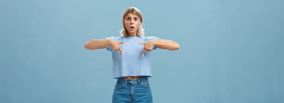Lifestyle. Studio shot of worried caucasian young woman feeling nervous her shoes do not fit frowning asking advice nervously pointing down shocked while frowning and standing over blue background.
