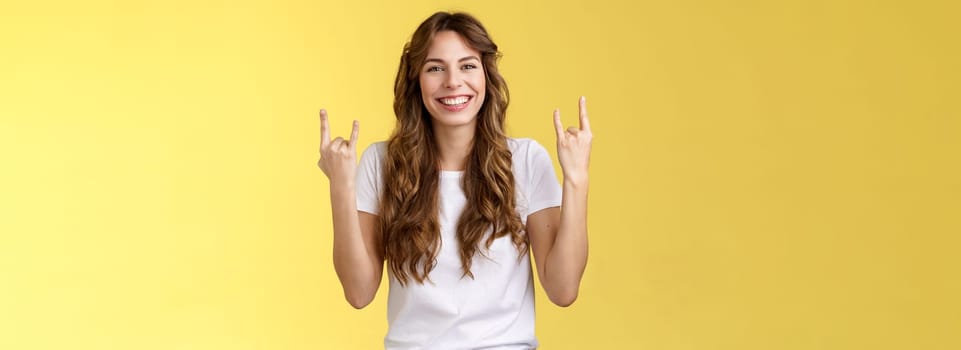 Happy cheerful feminine caucasian girl curly hairstyle show rock-n-roll heavy metal gesture smiling broadly enjoy awesome concert atmosphere stand yellow background joyfully cheering satisfied. Lifestyle.