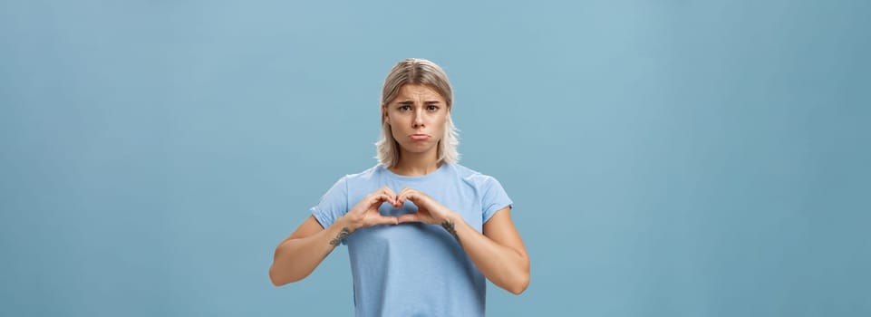 Heart being broken. Sad and gloomy heartbroken girl with blond hair tattoos on arms and tanned skin pursing lips whining and complaining making love sign over breast standing unhappy near blue wall.