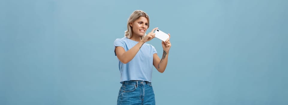 Girl trying to take photo of pet moving fast staring intense at smartphone screen pointing phone camera left feeling focused while taking picture on cellphone over blue background. Technology and lifestyle concept