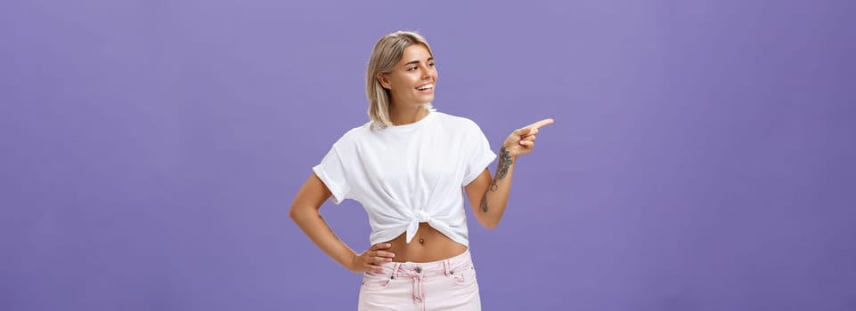 Hey nice job. Pleased carefree good-looking and confident female shop manager in stylish outfit holding hand on waist pointing and looking left with satisfied and delighted look over purple wall.