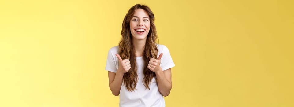 Pleased outgoing cheerful good-looking caucasian girl curly long haircut white t-shirt show thumbs up smiling lively pleased like awesome performance approve good choice yellow background. Lifestyle.