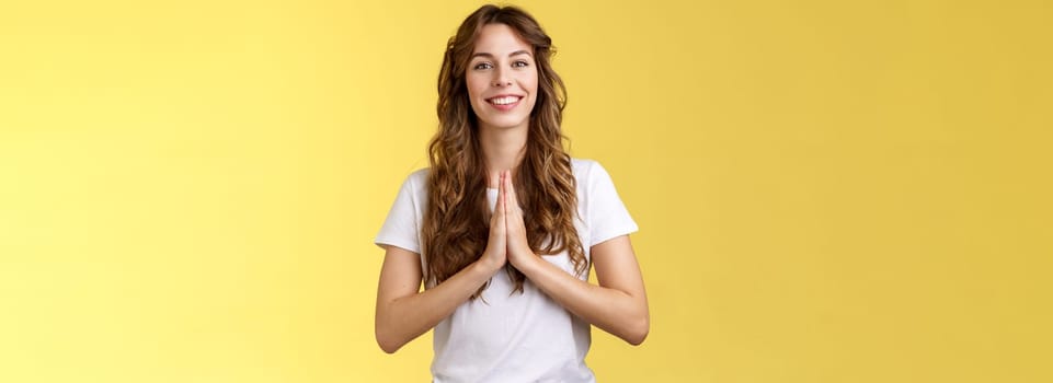 Cheerful confident outgoing young polite girl say namaste greeting asian guests smiling gladly thanking for visit hold hands pray supplication gesture asking favour begging stand yellow background. Lifestyle.