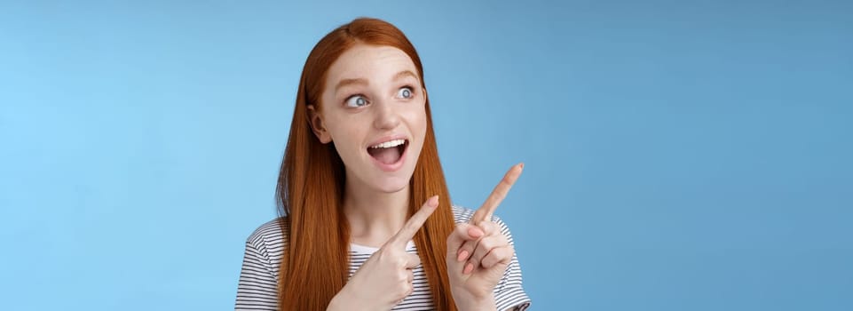 Lifestyle. Amused adorable redhead european girl say wow stare gasping astonished pointing looking upper left corner excited smiling drop jaw amazed standing thrilled blue background excitement concept.