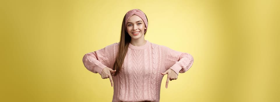 Tender young 20s european girl in sweater smiling broadly finally take-off braces pointing down promoting skillful dentist professional. Female student standing happy positive over yellow wall.