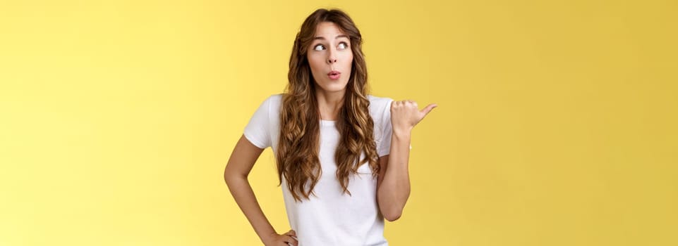 Girls gossiping discuss funny neighbours. Curious outgoing cute funny curly-haired woman look pointing thumb left fold lips wow amused glancing intrigued learn juicy rumor stand yellow background.