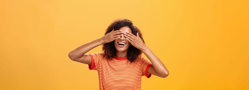Closing my eyes and counting ten. Portrait of charming dreamy and happy funny african american curly-haired female in striped trendy t-shirt covering sight with palm and smiling waiting surprise. Copy space