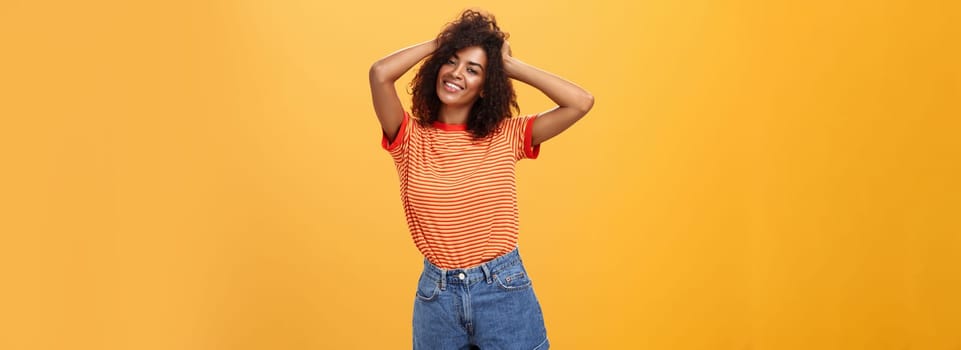 Girl feeling awesome making cool hairstyle posing near mirror feeling glad to have date tonight holding hands on curly hair smiling joyfully and tender standing against orange background. Lifestyle.