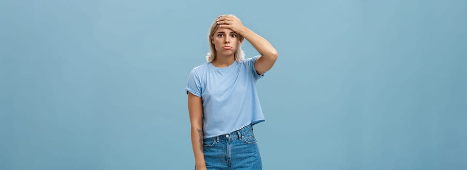 Lifestyle. Concerned and troubled young teenage girl being shocked with loads of responsibilities holding hand on forehead staring spaced out and pouting from tiresome perplexed situation over blue wall.