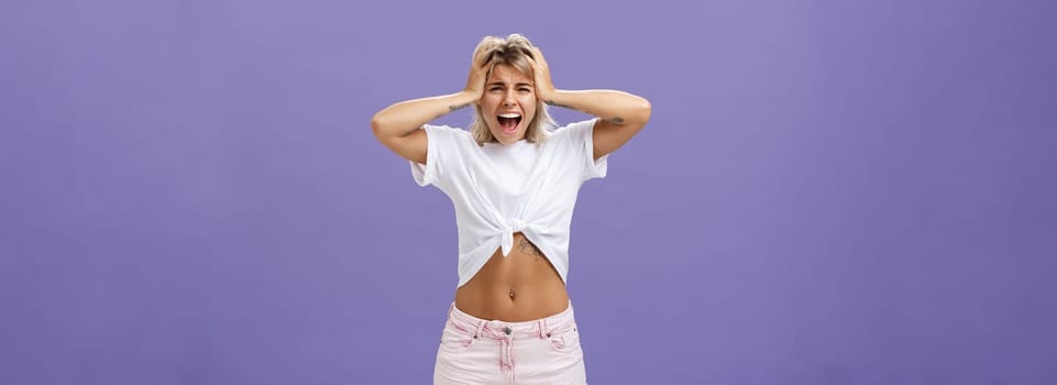 Lifestyle. Portrait of panicking concerned and depressed young european woman with blond hair tattoos and athletic body yelling out loud holding hands on head being concerned and shook over purple background.