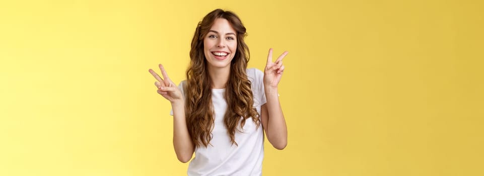 Friendly peaceful charming happy young girl curly long hair show peace victory signs having fun posing outgoing lively look camera amused laughing smiling broadly stand yellow background. Lifestyle.