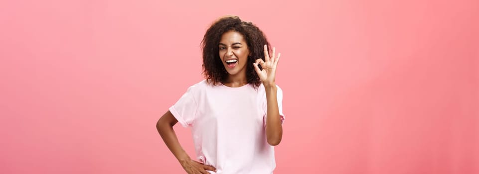 Girl got us covered. Charming charismatic and confident african american skillful woman in stylish outfit holding hand on waist showing okay gesture and winking assured at camera over pink wall.