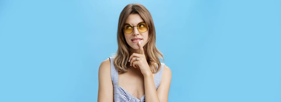 Flirty and creative charming feminine woman in stylish sunglasses and top holding finger on lip gazing at upper right corner thoughtful making up plan or thinking about tasty cake she wants. Body language concept