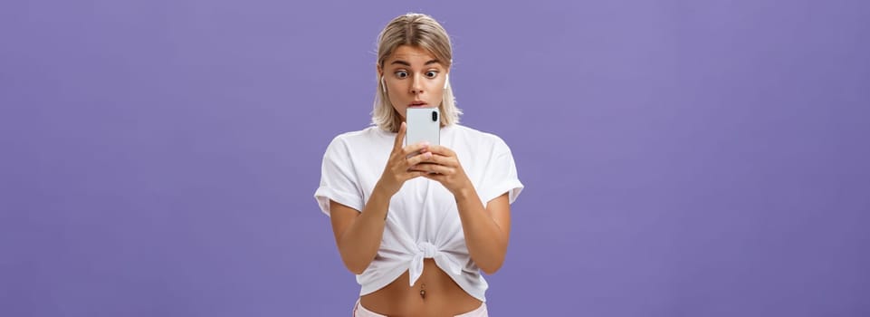 Girl watching thrilling video on social media in wireless earphones holding smartphone near face popping eyes at smartphone screen gazing at gadget with interest and curiosity over purple background. Technology concept