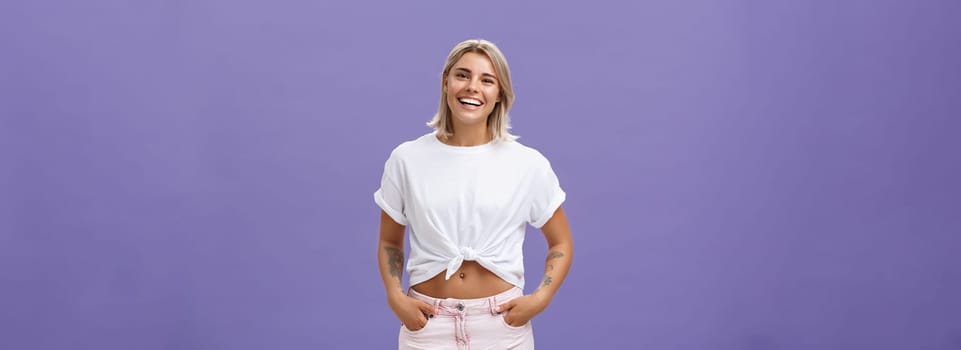 Lifestyle. Joyful attractive tanned girlfriend with blond hair and tattoos on arms with cropped top chilling by pool with friends talking with amused happy expression smiling at camera holding hands in pockets.