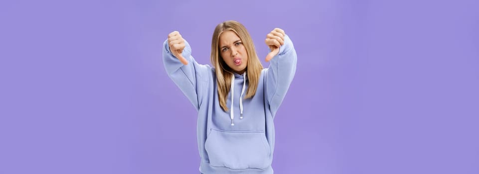 Indoor shot of moodie displeased and unimpressed cool modern female with blond hair and tanned skin tilting head sticking out tongue showing thumbs down in dissatisfaction posing against purple wall. Body language concept