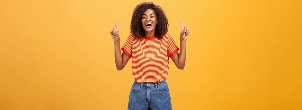 Woman feeling amused and entertained. Portrait of happy carefree stylish African-American girl with afro hairstyle laughing out loud joyfully pointing up with raised arms over orange wall. Emotions concept