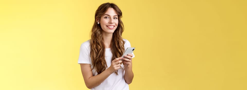 Feminine lovely curly-haired woman white t-shirt hold smartphone picked awesome new song listen music wearing wireless earphones smiling delighted camera enjoy earbuds beats yellow background. Lifestyle.