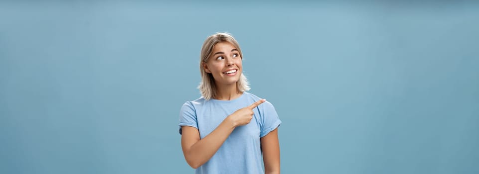 Lifestyle. Waist-up shot of creative and dreamy happy attractive blonde female in casual t-shirt looking and pointing at upper left corner with admiration and joy standing over blue background amused and curious.