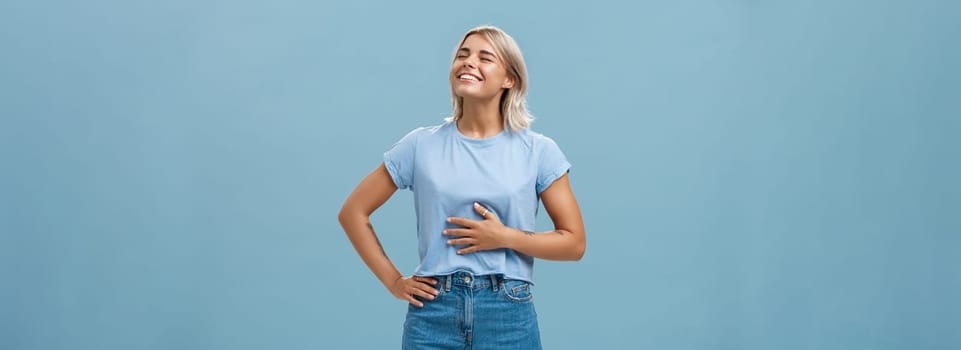 Cute girl likes eat after workout being pleased and stuffed after dinner. Portrait of satisfied attractive blonde young woman touching belly raising head up with broad smile over blue background.