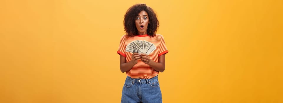 Woman shocked finding lots of cash in safe. Portrait of surprised speechless good-looking dark-skinned female with curly haircut folding lips gasping holding money posing over orange background.