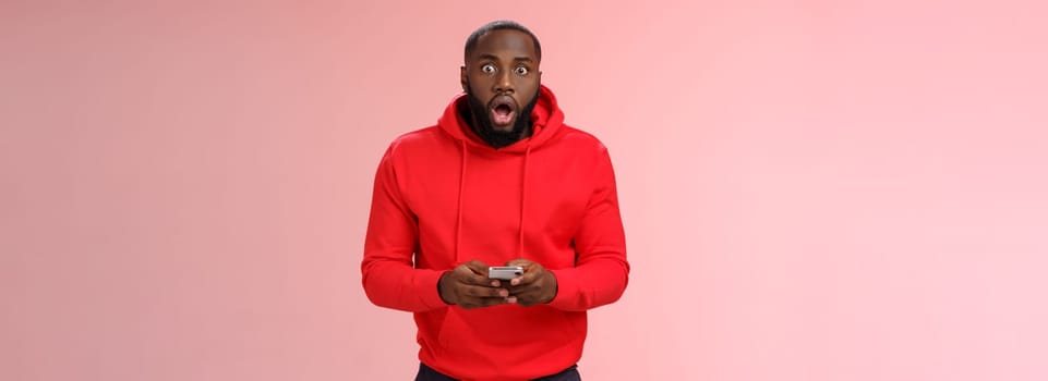 Amused overhwelmed black bearded guy in red hoodie drop jaw gasping widen eyes staring camera impressed holding smartphone using amazing cool game astonished new phone features, standing pink wall.